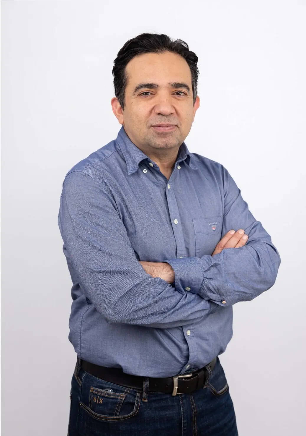 Dr Reza Sabry, General Practitioner, stands with arms crossed in blue shirt and jeans on a white background.