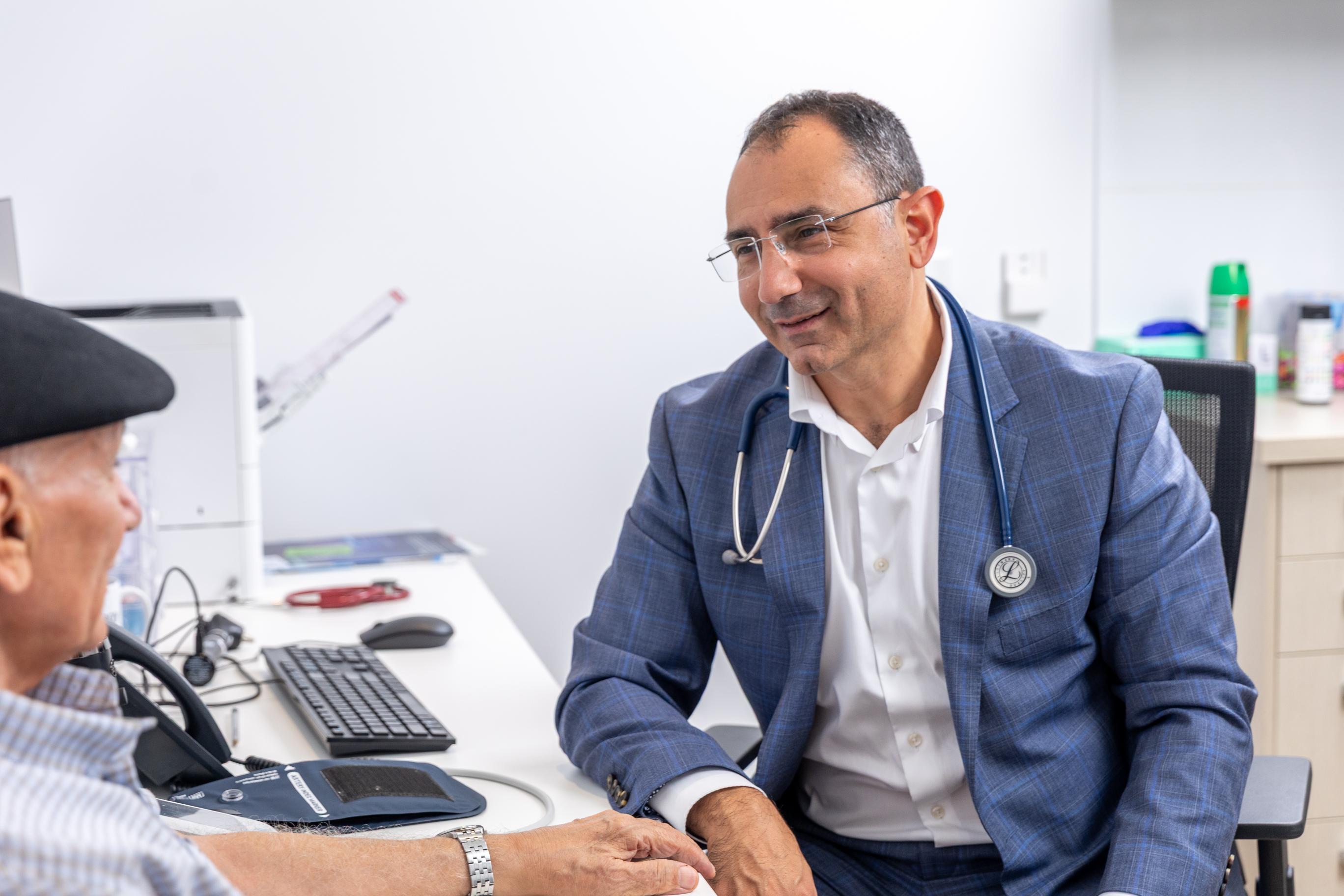 Dr Nadim in suit with a stethoscope consults with an elderly man in a clinic.