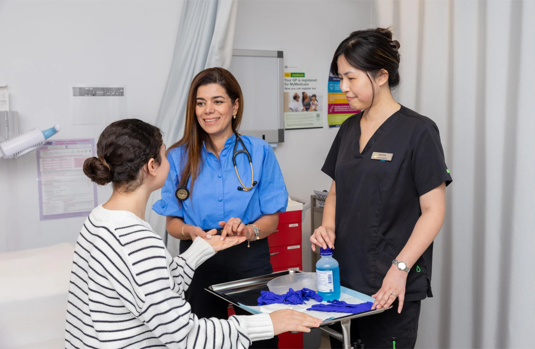Dr Neda with a stethoscope smiling at a patient while discussing at a clinic counter, a nurse stands nearby with a tray of gloves and supplies.