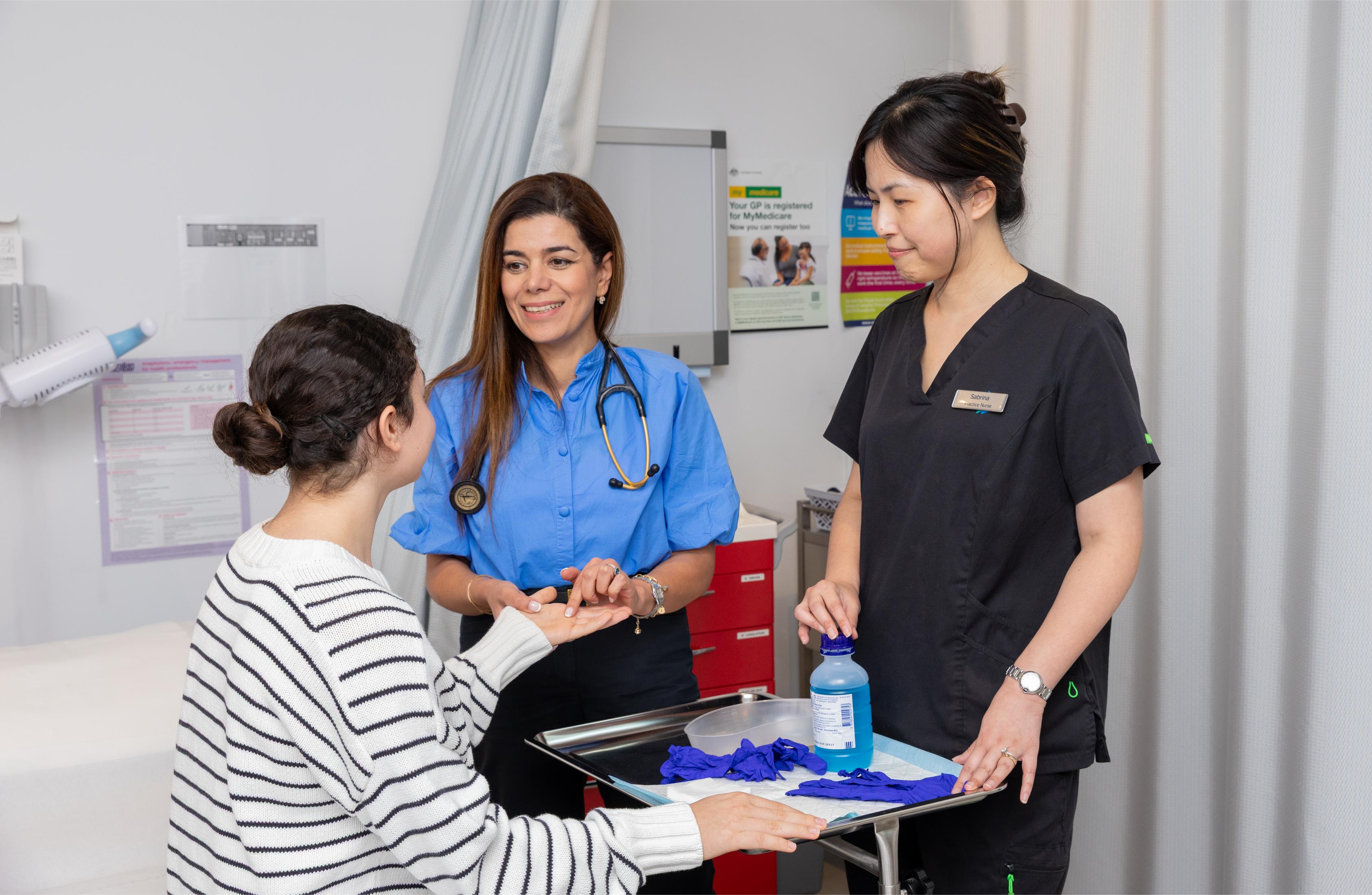 Dr Neda with a stethoscope smiling at a patient while discussing at a clinic counter, a nurse stands nearby with a tray of gloves and supplies.