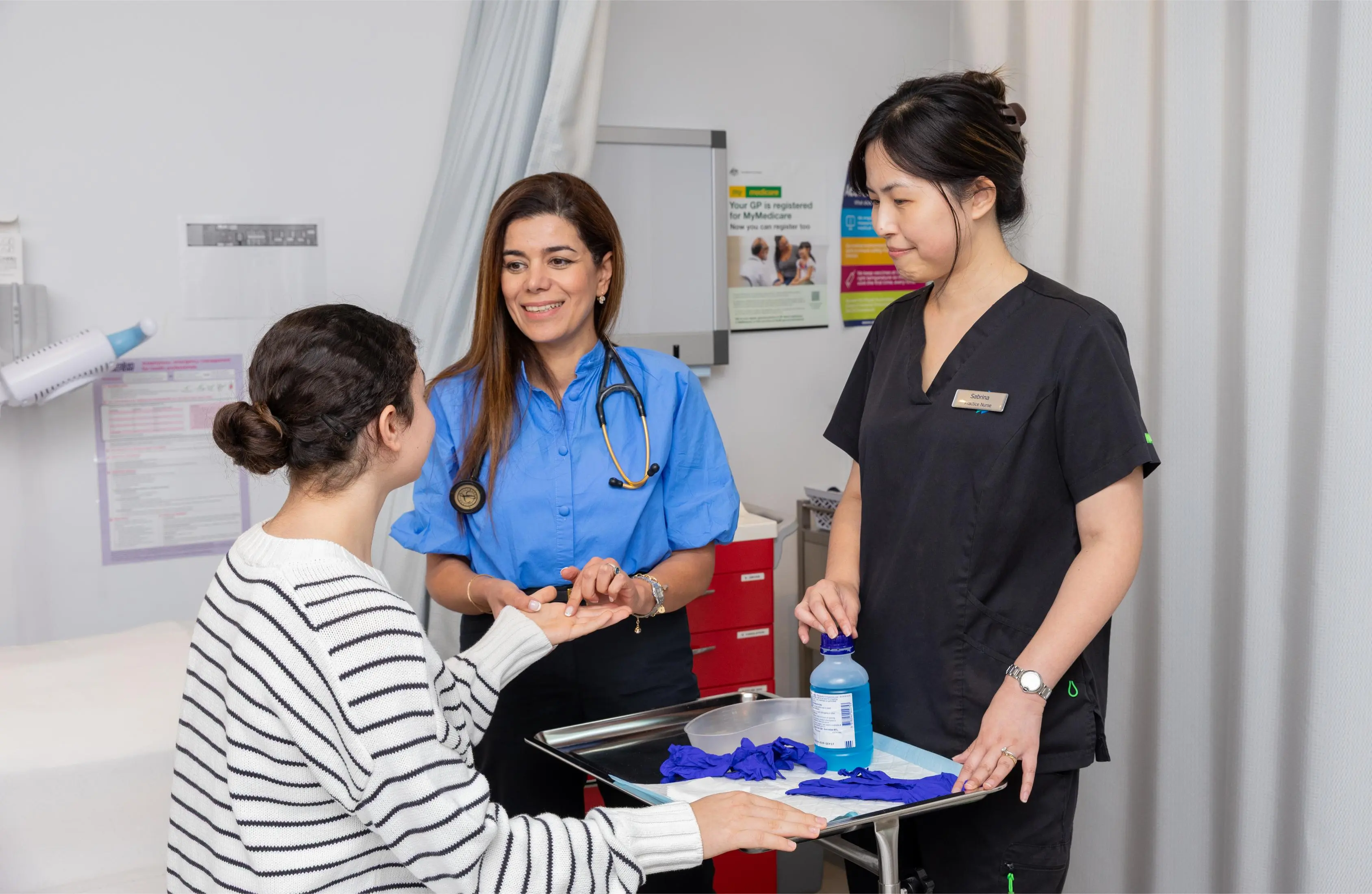 Dr Neda with a stethoscope smiling at a patient while discussing at a clinic counter, a nurse stands nearby with a tray of gloves and supplies.