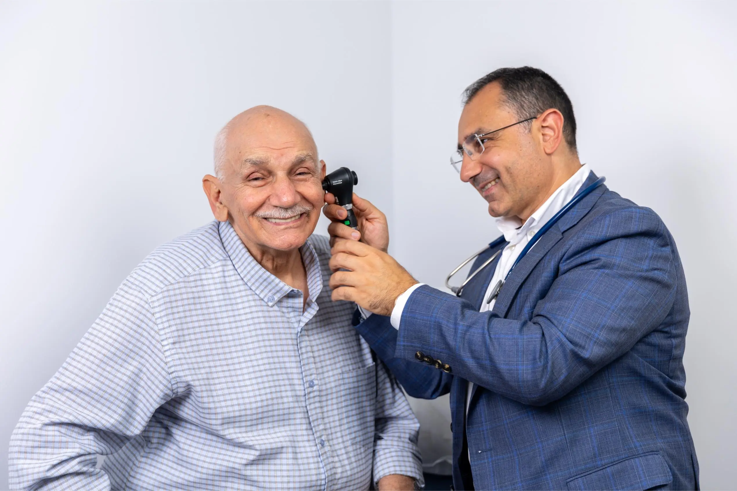 Dr Nadim in a blue suit examines an elderly man's ear with an otoscope during a medical checkup, both smiling.