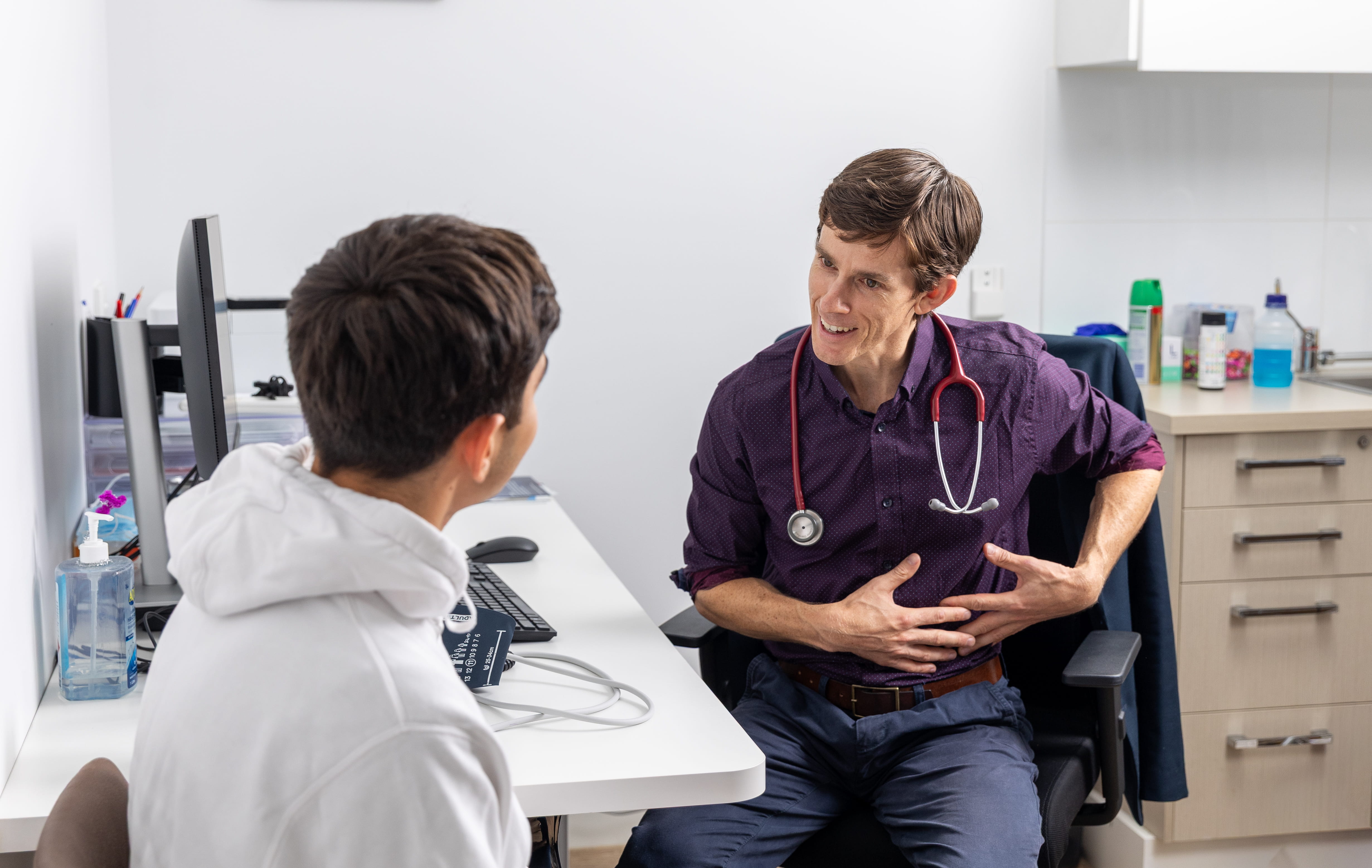 Dr Casey with stethoscope consulting a patient at a clinic desk, discussing health.