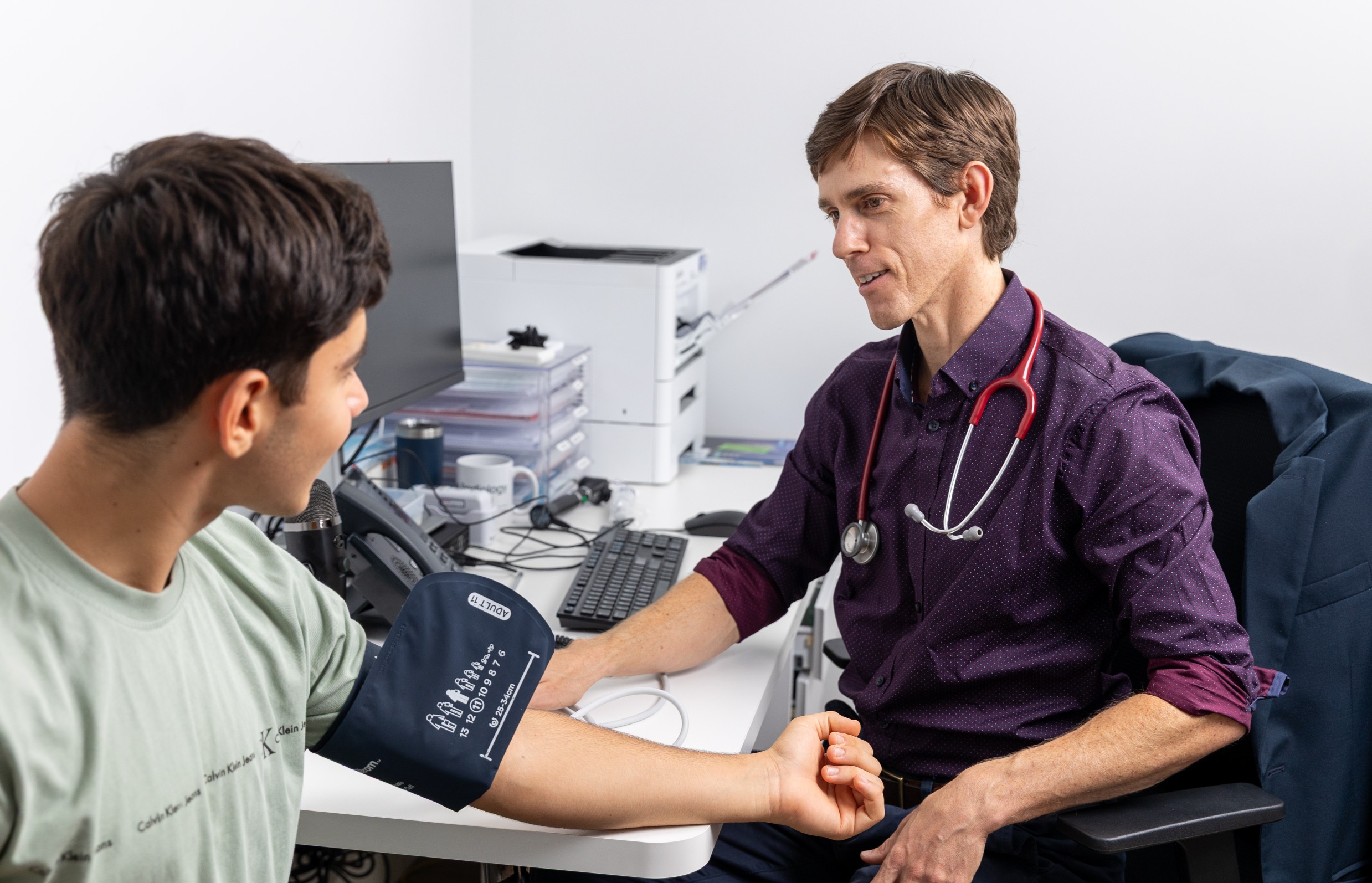Dr Casey in a purple shirt checks a patient's blood pressure with a cuff and stethoscope in a clinic setting