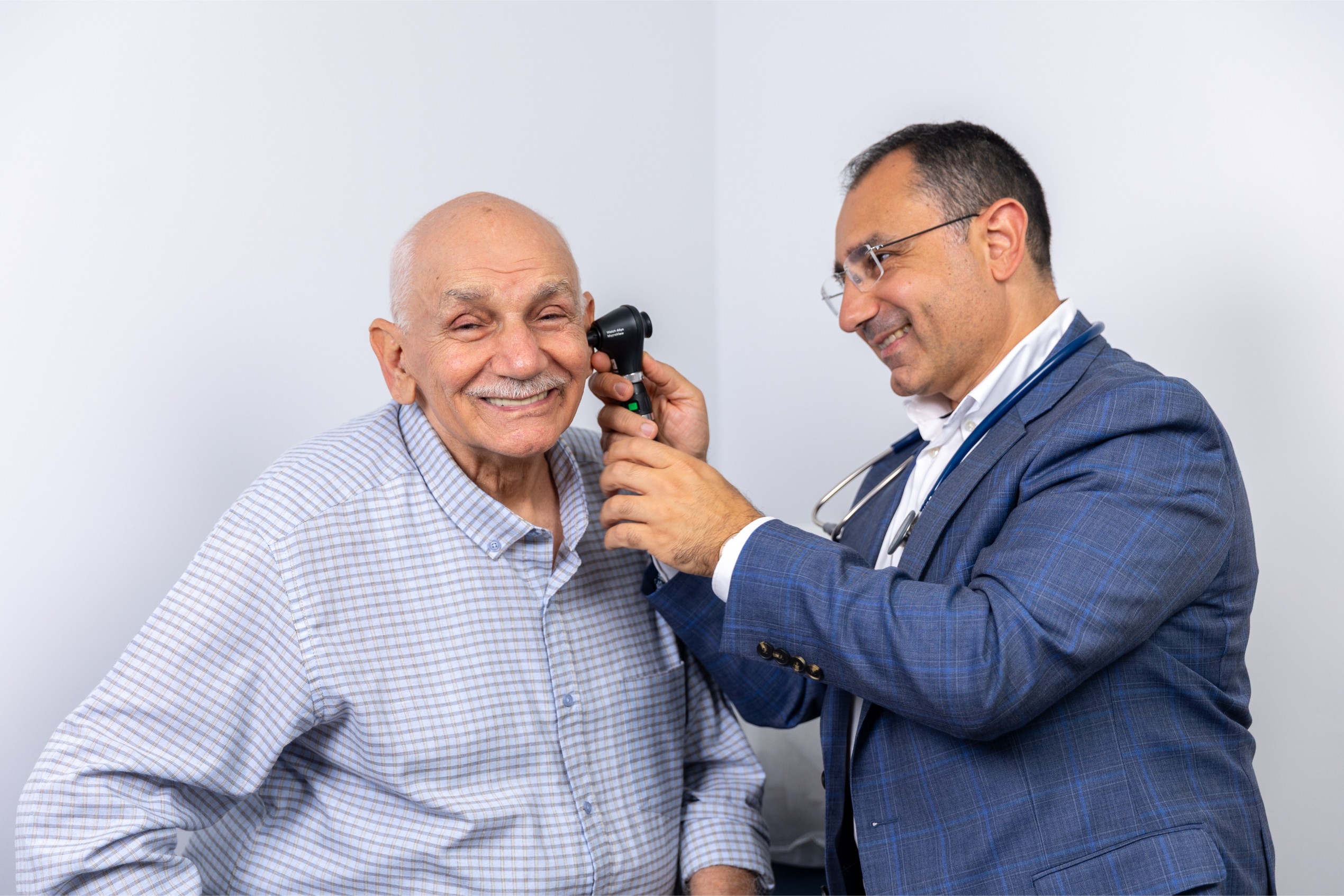 Dr Nadim examines an elderly man's ear using an otoscope in a clinical setting.