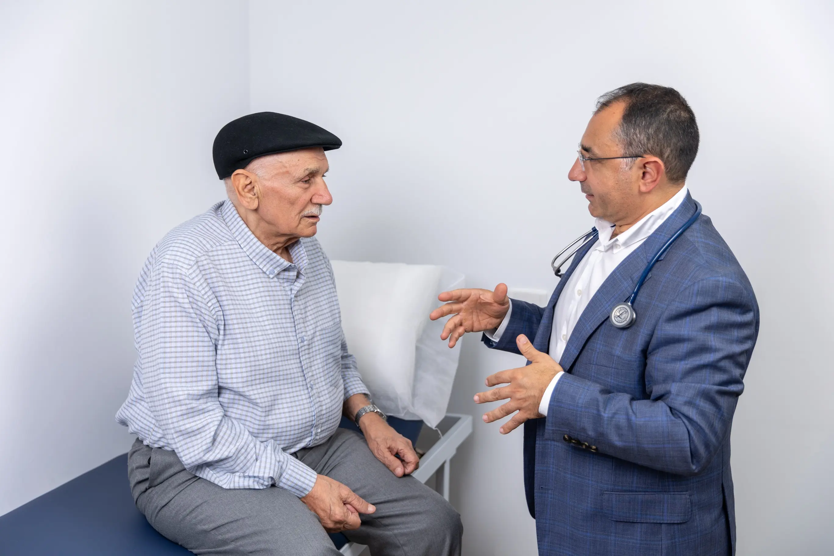 Dr Nadim discusses Men’s Health with an older man in a black flat cap sitting on an exam table.