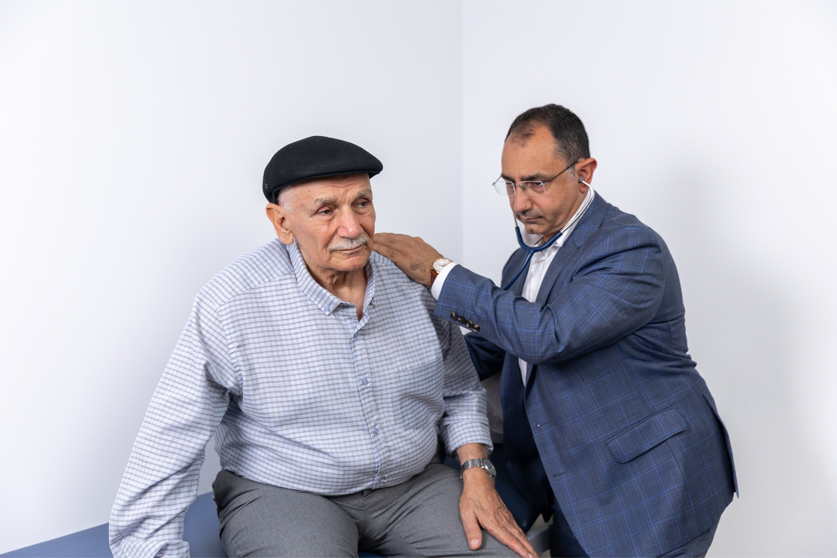 Dr Nadim in a blue suit listens with a stethoscope to an elderly man wearing a checkered shirt and a black cap during a medical exam.