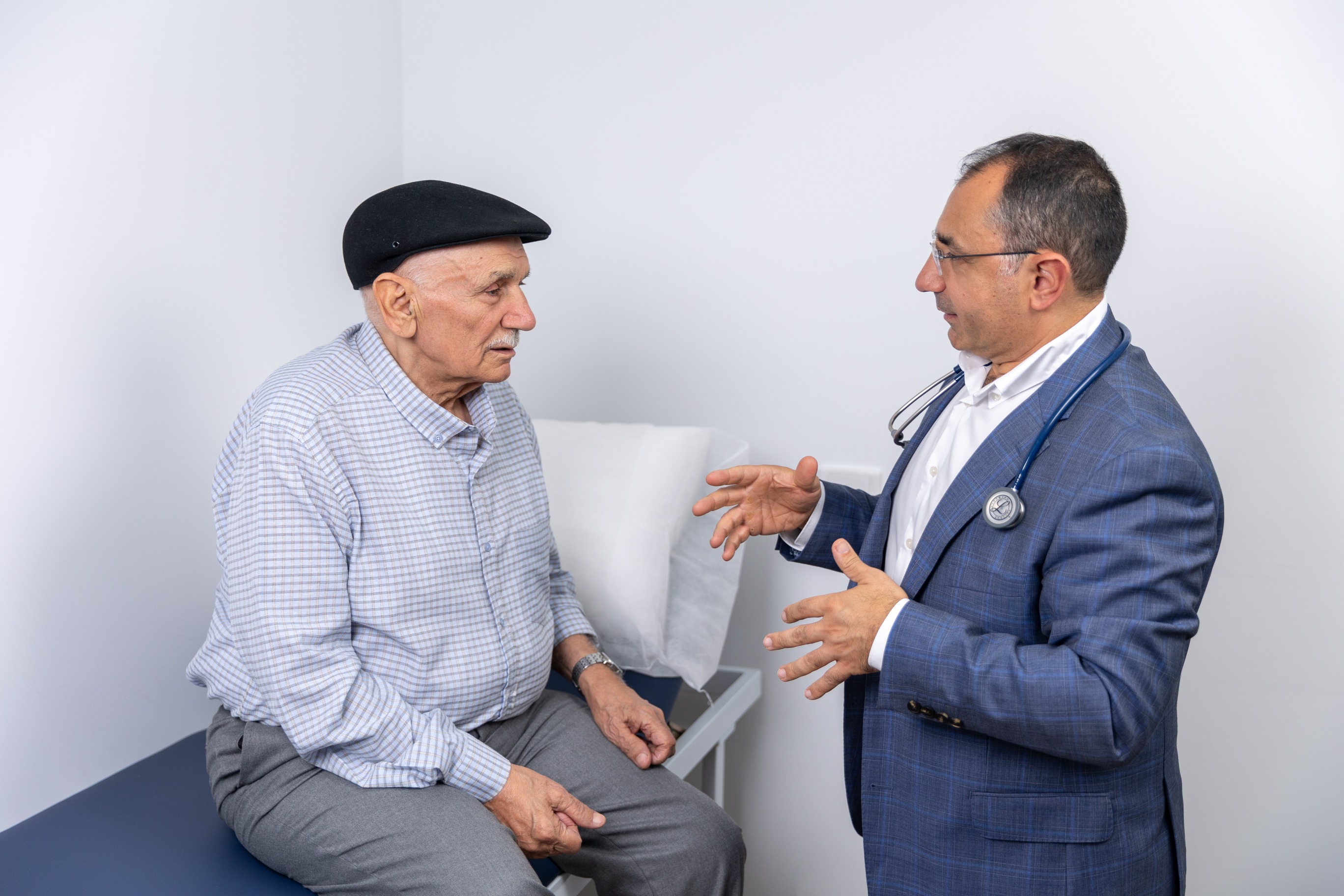 Dr Nadim discusses travel health and vaccines with an older man on an exam table in a medical office.