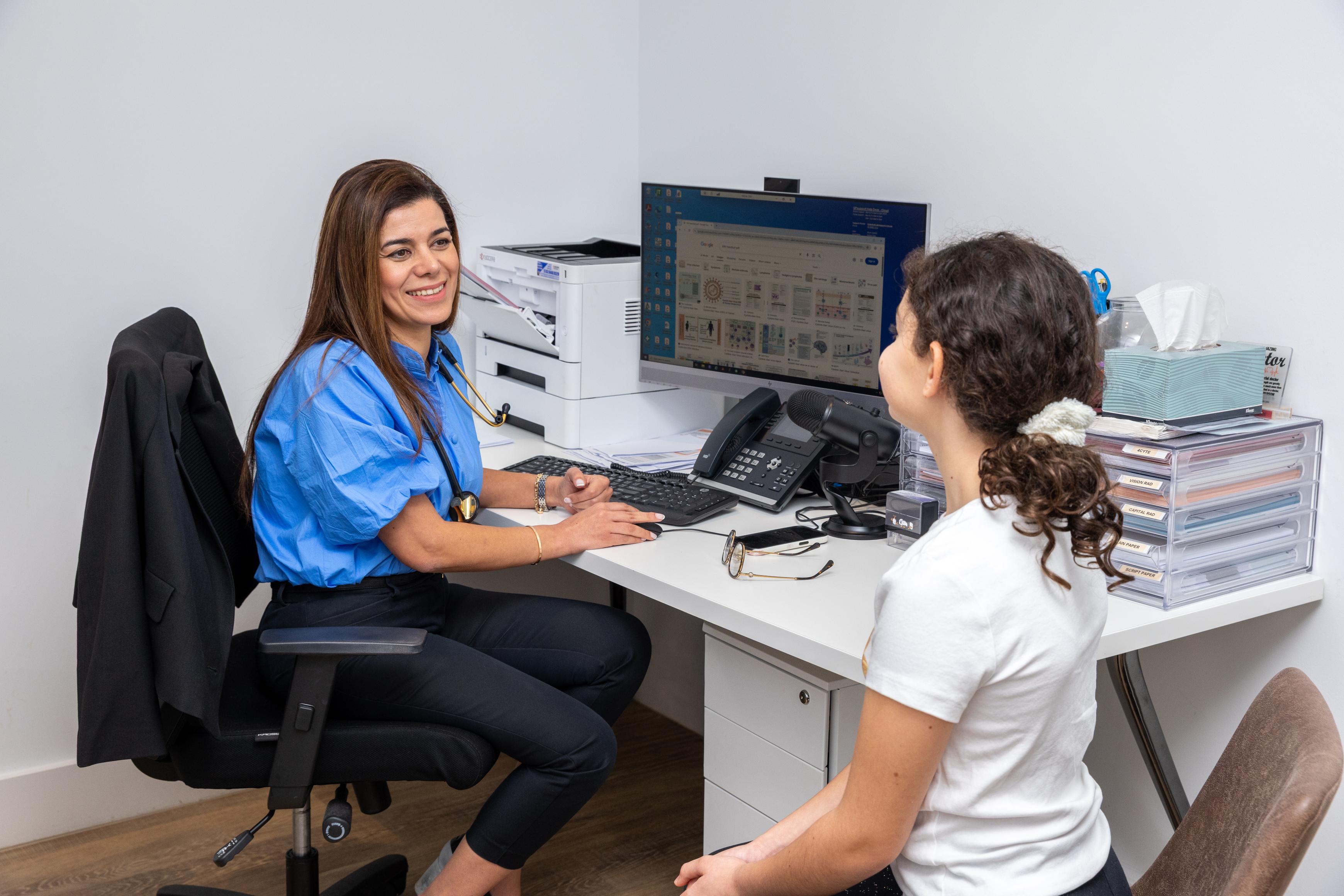 Dr Neda in a blue shirt discusses iron deficiency management with a child at her desk, office supplies and monitor nearby.