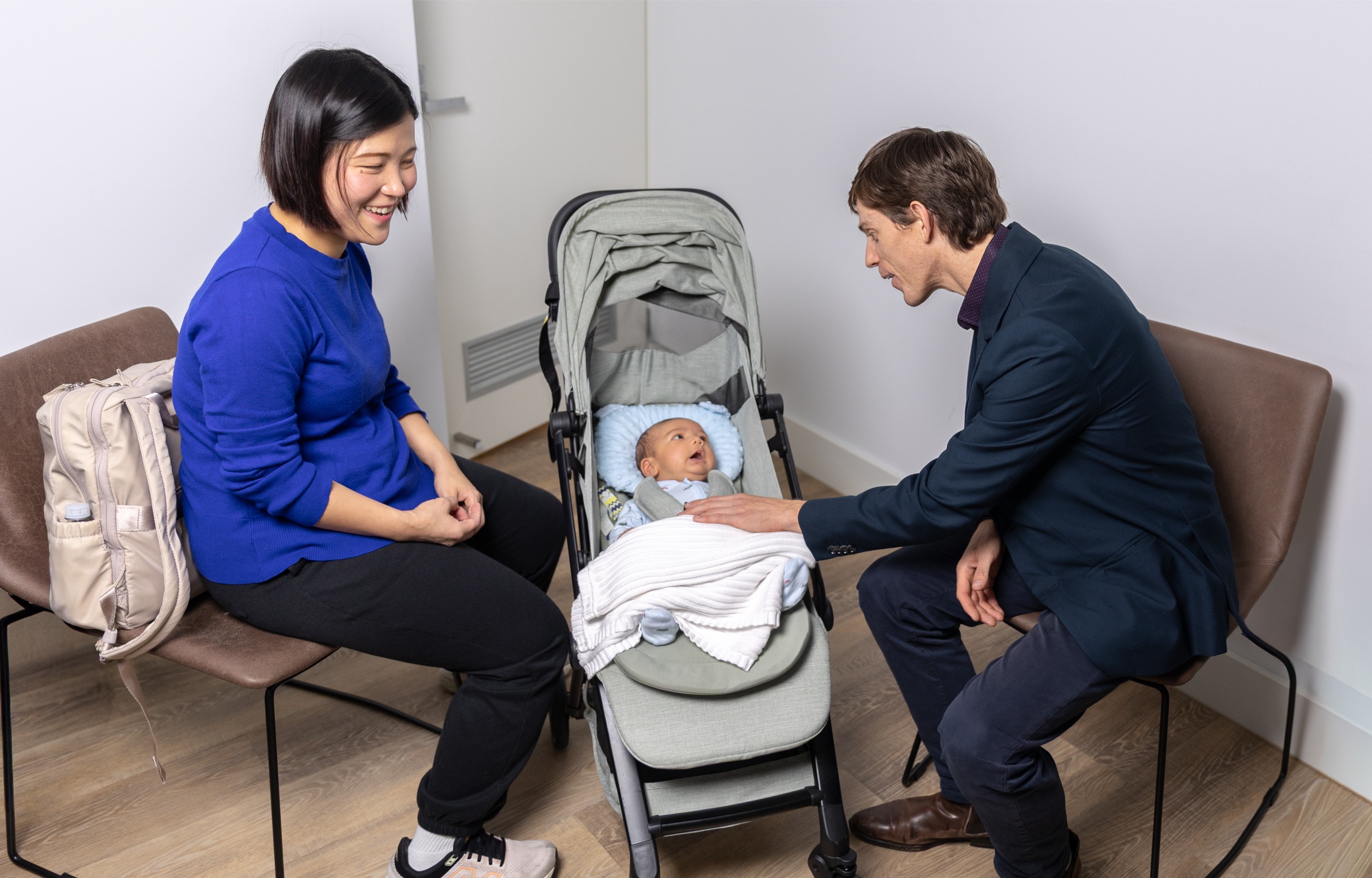Calm setting for paediatric & adult immunisation with baby in stroller, woman beside, man touching baby’s arm indoors.
