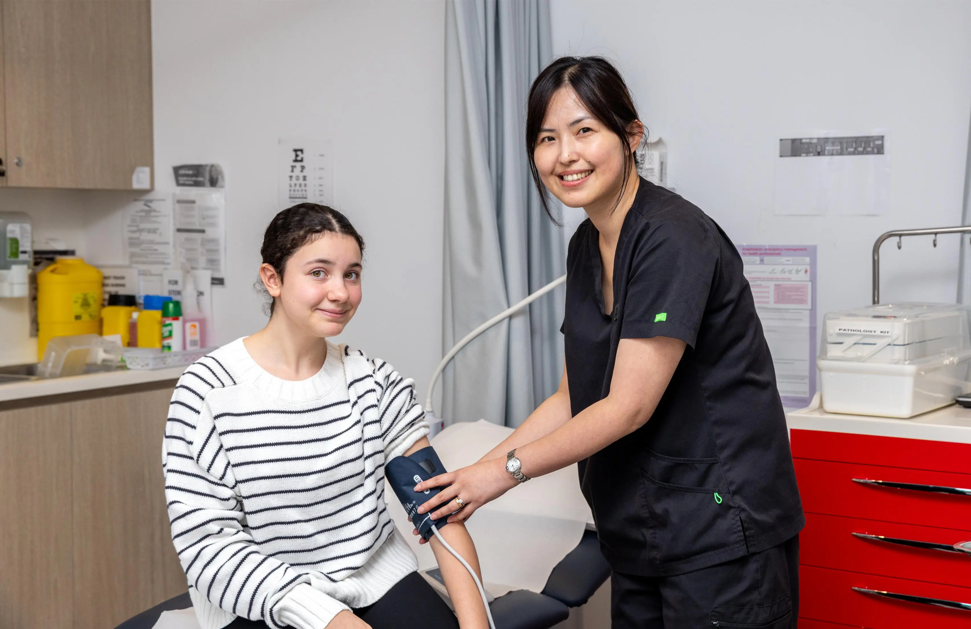 Healthcare professional in dark scrubs checks a patient’s blood pressure in a clinical room; patient sits smiling wearing a striped sweater.