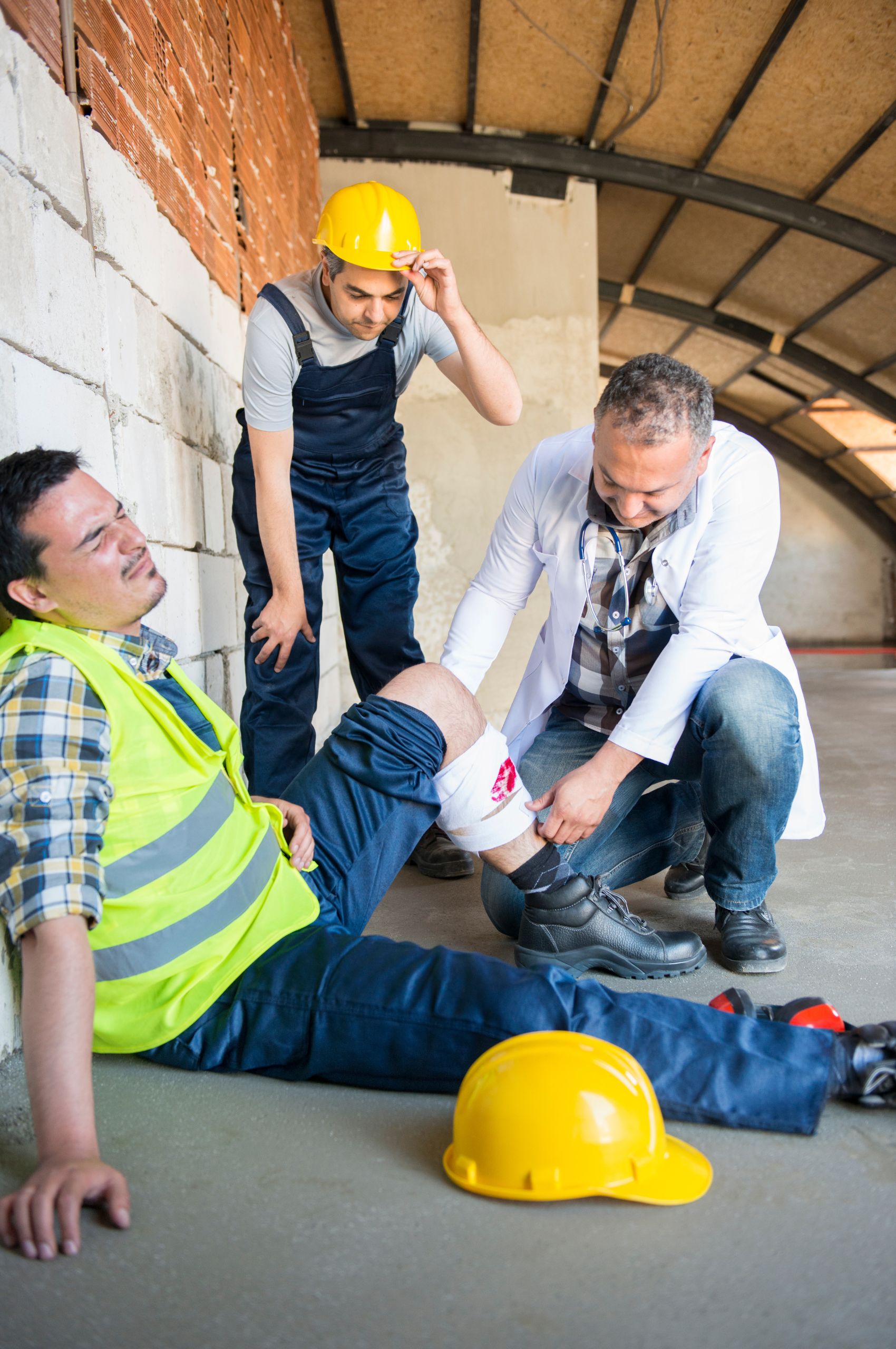 Doctor bandages a worker’s injured knee under Workcover & TAC; another worker watches, safety vests and helmets visible.
